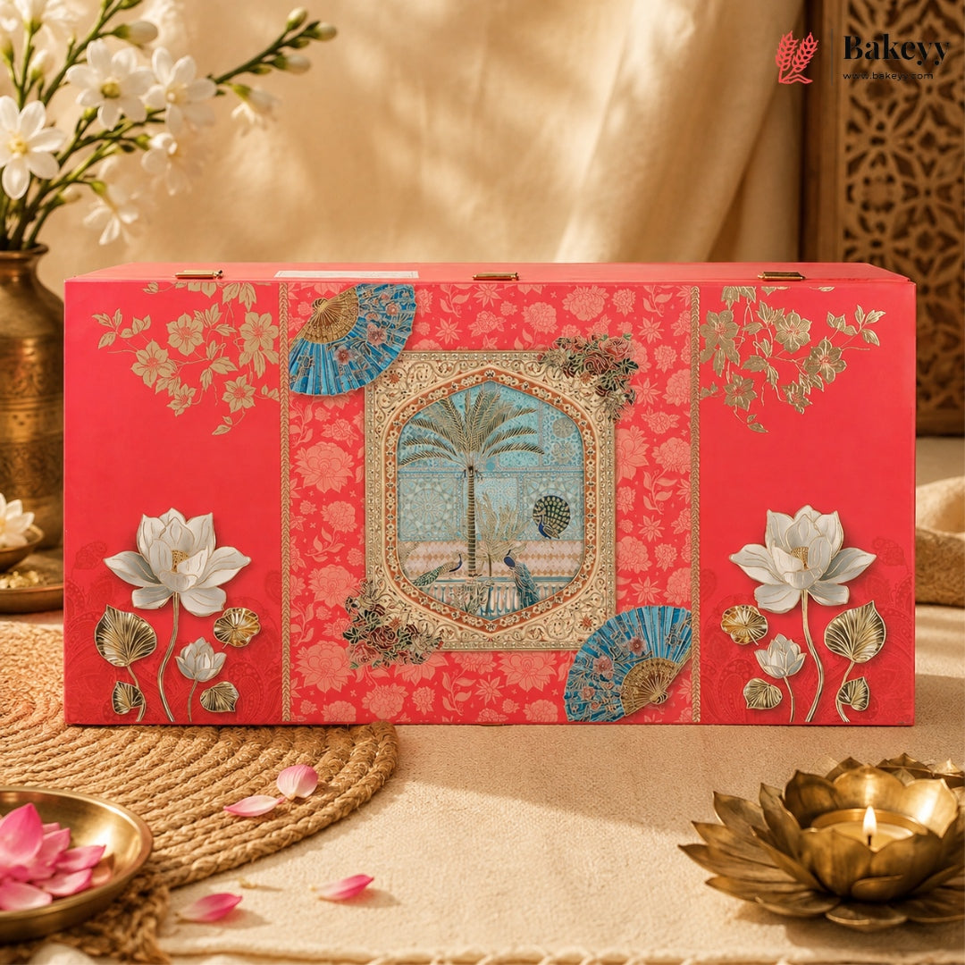 A decorative red gift box with intricate gold floral patterns, blue fan motifs, and a central illustration of a palm tree and peacock, styled on a beige fabric surface with floral props and a candle.