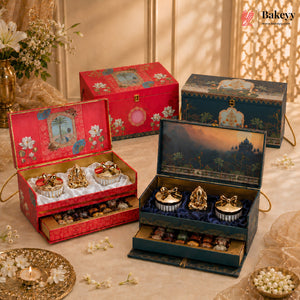 Three ornate decorative gift boxes for chocolates are arranged on a draped beige fabric with floral props. One red box is open in the foreground showing two chocolate cups and chocolates inside, one dark blue box is open showing two chocolate cups and chocolates inside, and one closed red box stands behind.