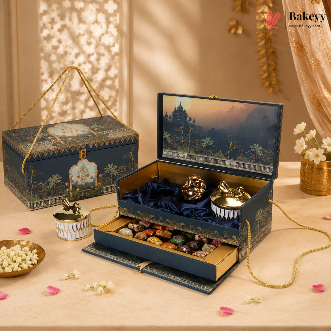 An ornate dark blue gift box set with gold detailing, shown open to reveal a jewelry tray, decorative items, and a lower drawer with assorted chocolates or sweets. A matching closed box sits beside it on a beige surface with warm decorative props and shadows.
