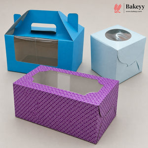 Three paper bakery boxes on a light beige background: a blue handled box with a clear window, a light blue cube with a round clear window, and a purple patterned rectangular box with an octagonal clear window. A Bakeyy logo appears in the top right corner.