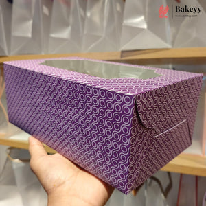 A hand holding a purple bakery box with a repeating circular pattern and a clear window on top, set against a background of white textured bags and wooden shelves.