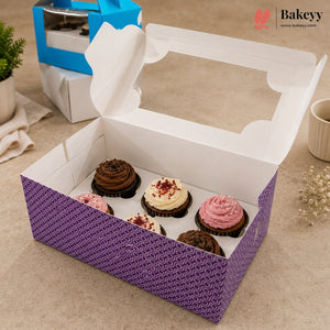 An open purple bakery box with a clear window lid, holding six decorated cupcakes, on a beige tabletop. A blue and white box sits in the background, with a small plant and cup visible around it.