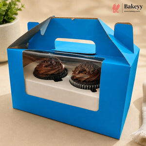 A bright blue cardboard cupcake carrier box with a clear plastic window, shown holding two chocolate frosted cupcakes. The box has a built-in handle and is photographed on a beige tabletop with a small potted plant in the background.