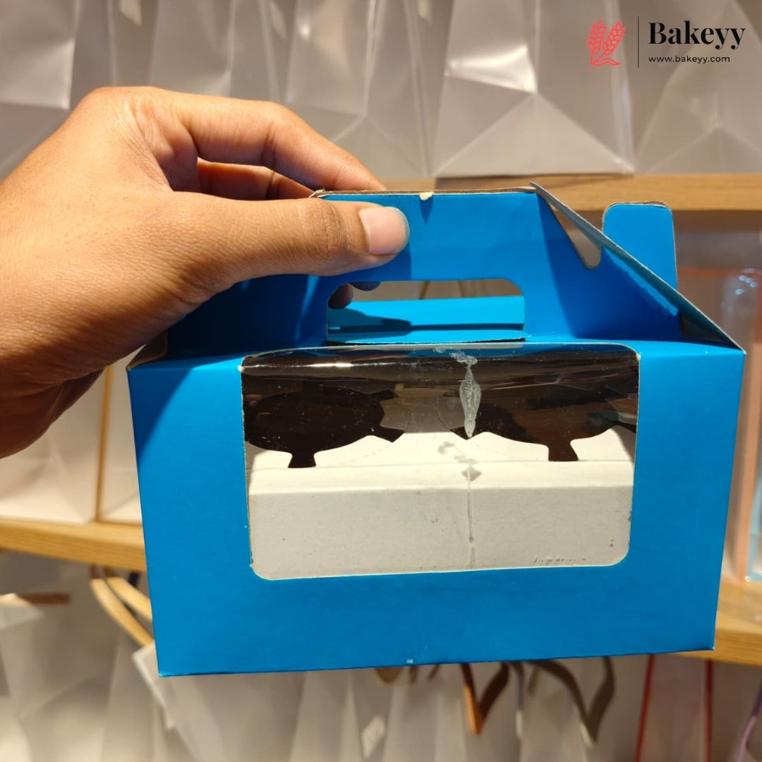 A hand holding a bright blue bakery box with a clear plastic window showing a white tray inside, set against a blurred background with a Bakeyy logo in the top right corner.