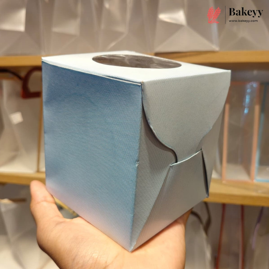 A hand holding a light blue and white textured square box with a circular cutout on top, set against a blurred bakery shelf background with a Bakeyy watermark.