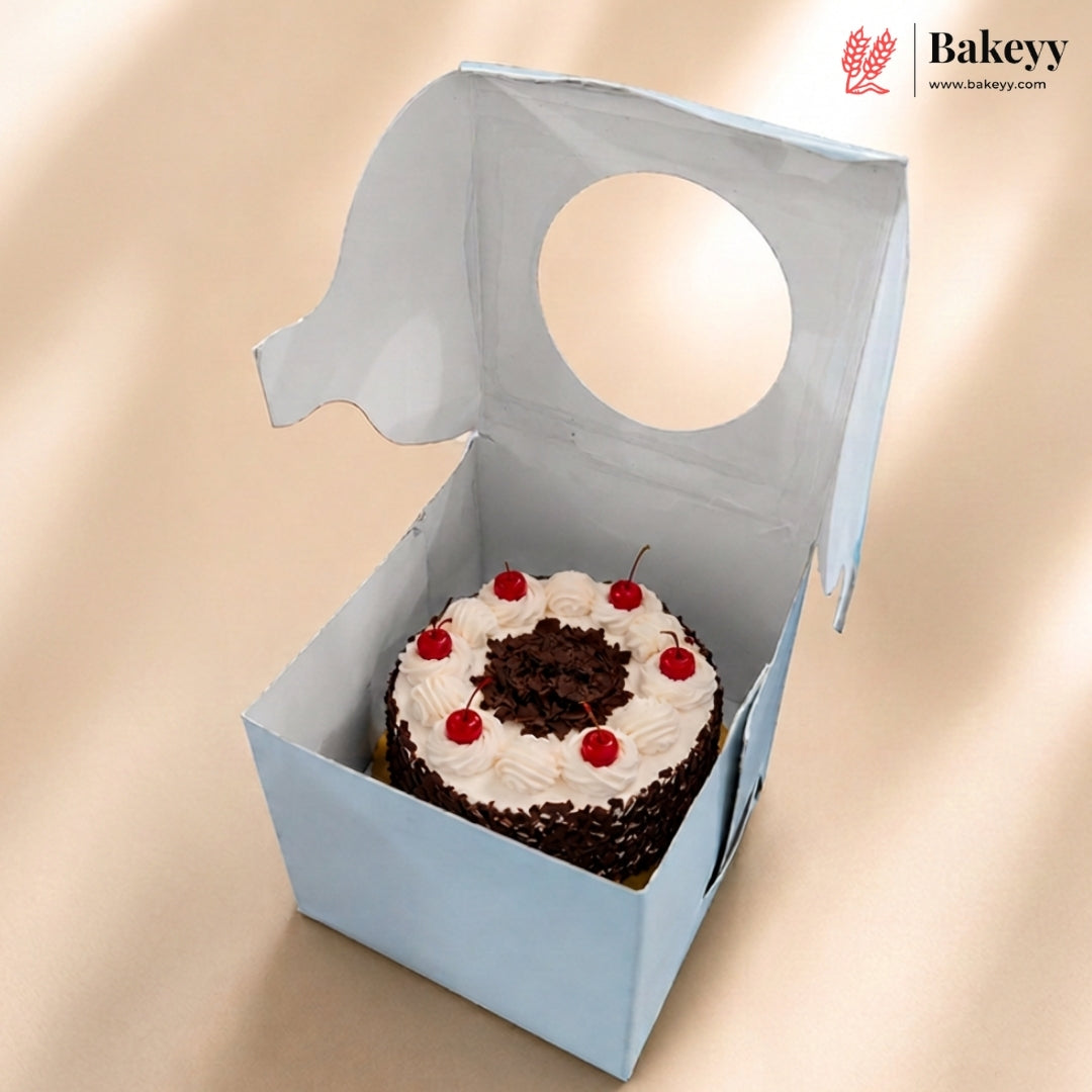 A small round chocolate cake with white frosting and red cherries inside a light blue bakery box with a circular cutout, shown on a beige background. The Bakeyy logo is visible in the top right corner.