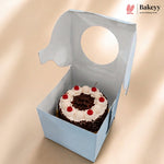 A small round chocolate cake with white frosting and red cherries inside a light blue bakery box with a circular cutout, shown on a beige background. The Bakeyy logo is visible in the top right corner.