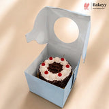 A small round chocolate cake with white frosting and red cherries inside a light blue bakery box with a circular cutout, shown on a beige background. The Bakeyy logo is visible in the top right corner.