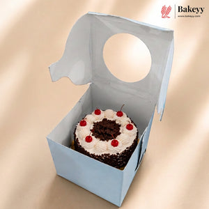 A small round chocolate cake with white frosting and red cherries inside a light blue bakery box with a circular cutout, shown on a beige background. The Bakeyy logo is visible in the top right corner.
