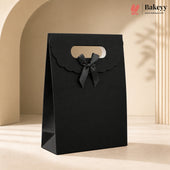 A black paper gift bag with scalloped handle edges and a matching black ribbon bow, standing on a beige surface in a softly lit studio setting. The Bakeyy logo appears in the top right corner.