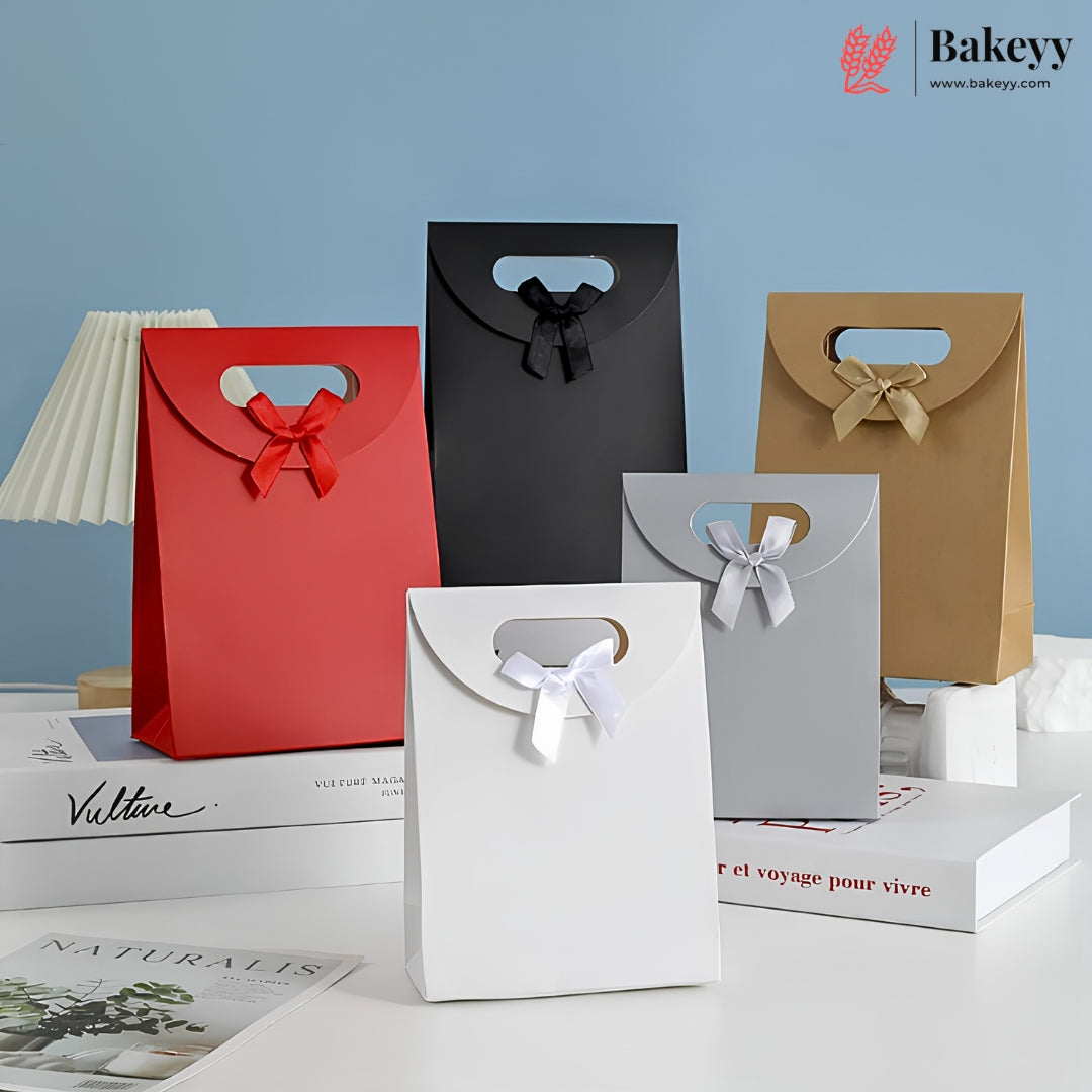 Five paper gift bags in red, black, brown, white, and gray, each with a handle cutout and a ribbon bow at the top, arranged on books against a light blue background. The Bakeyy logo appears in the top right corner.