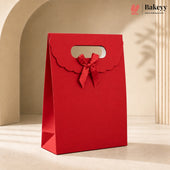 A red paper gift bag with a scalloped handle and a matching red ribbon bow, standing on a beige surface in a softly lit studio setting. The Bakeyy logo appears in the top right corner.