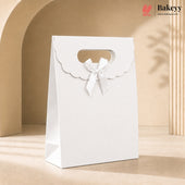 A white bakery gift bag with a scalloped handle cutout and a white satin bow, standing on a beige surface against a warm beige background with soft shadows. The Bakeyy logo is visible in the top right corner.