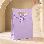 A lavender purple paper gift bag with a scalloped handle and a matching ribbon bow, standing on a beige surface against a warm beige background with soft shadows.