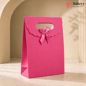 A bright pink paper gift bag with scalloped handle cutouts and a matching pink ribbon bow, standing on a beige surface in a softly lit studio setting. The Bakeyy logo appears in the top right corner.