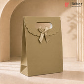 A kraft paper bakery gift bag in a warm beige-tan color, featuring a scalloped handle cutout and a matching satin ribbon bow. The bag is photographed in a warm, minimalist studio setting with soft shadows.