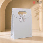 A tall white bakery gift bag with a scalloped flap detail and a white satin bow, standing on a beige surface against a warm beige background with soft shadows.
