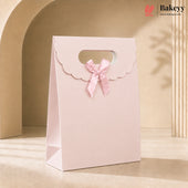 A light pink paper gift bag with a scalloped flap handle, a matching pink ribbon bow, and a cutout window near the top, photographed in a warm beige studio setting with soft shadows. The Bakeyy logo appears in the top right corner.