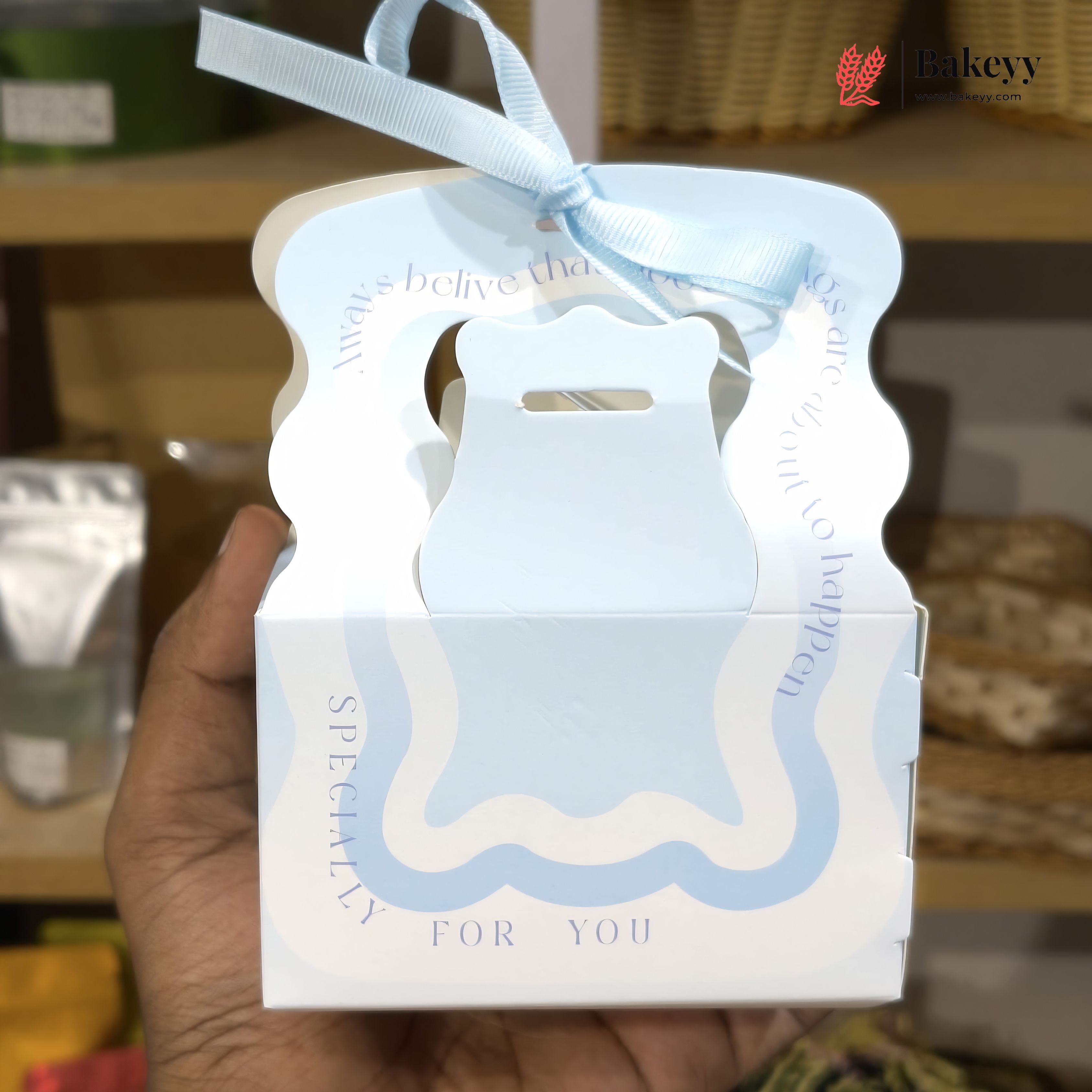 Hand holding a white and blue gift box with a bear design, featuring the brand 'Bakeyy'.