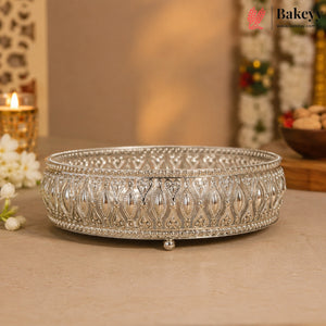 An ornate silver-colored metal tray with intricate filigree patterns and small feet, displayed on a beige surface with a warm, softly blurred background.
