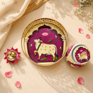 A decorative round tray and matching small container with a hand-painted style illustration of a white cow under a purple arch, styled on a beige surface with pink petals and a gold-toned lamp.