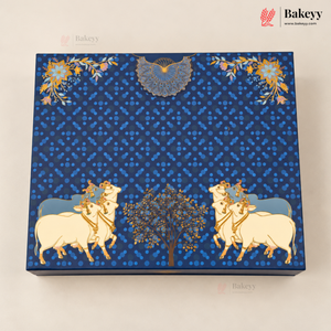 A square blue decorative box with intricate gold and white illustrations of cows, a tree, floral motifs, and a bird, photographed on a light beige background. The Bakeyy logo is visible in the top right corner.