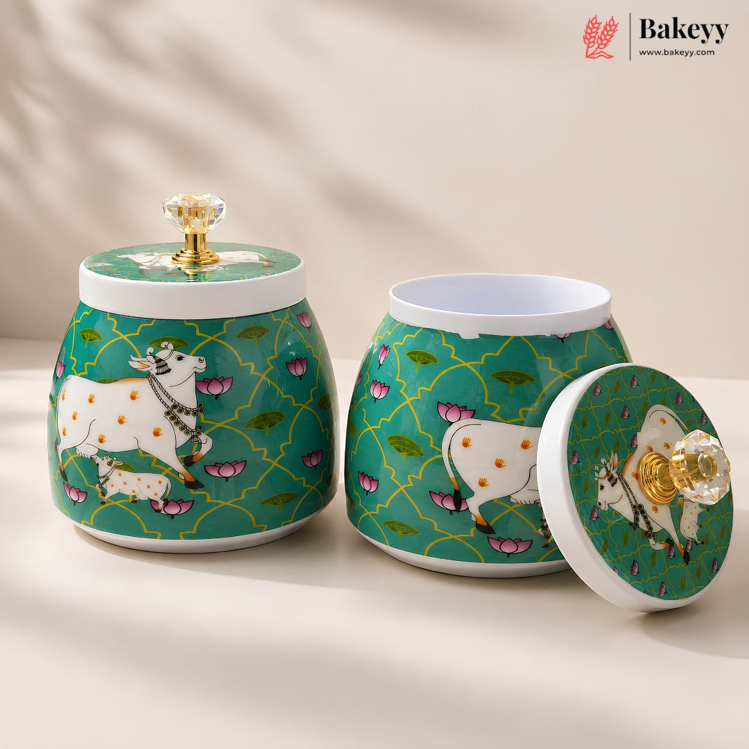 Two decorative ceramic canisters with matching lids on a warm beige surface. The canisters have a teal green background with a gold trellis pattern, pink lotus motifs, and white cows with yellow spots.