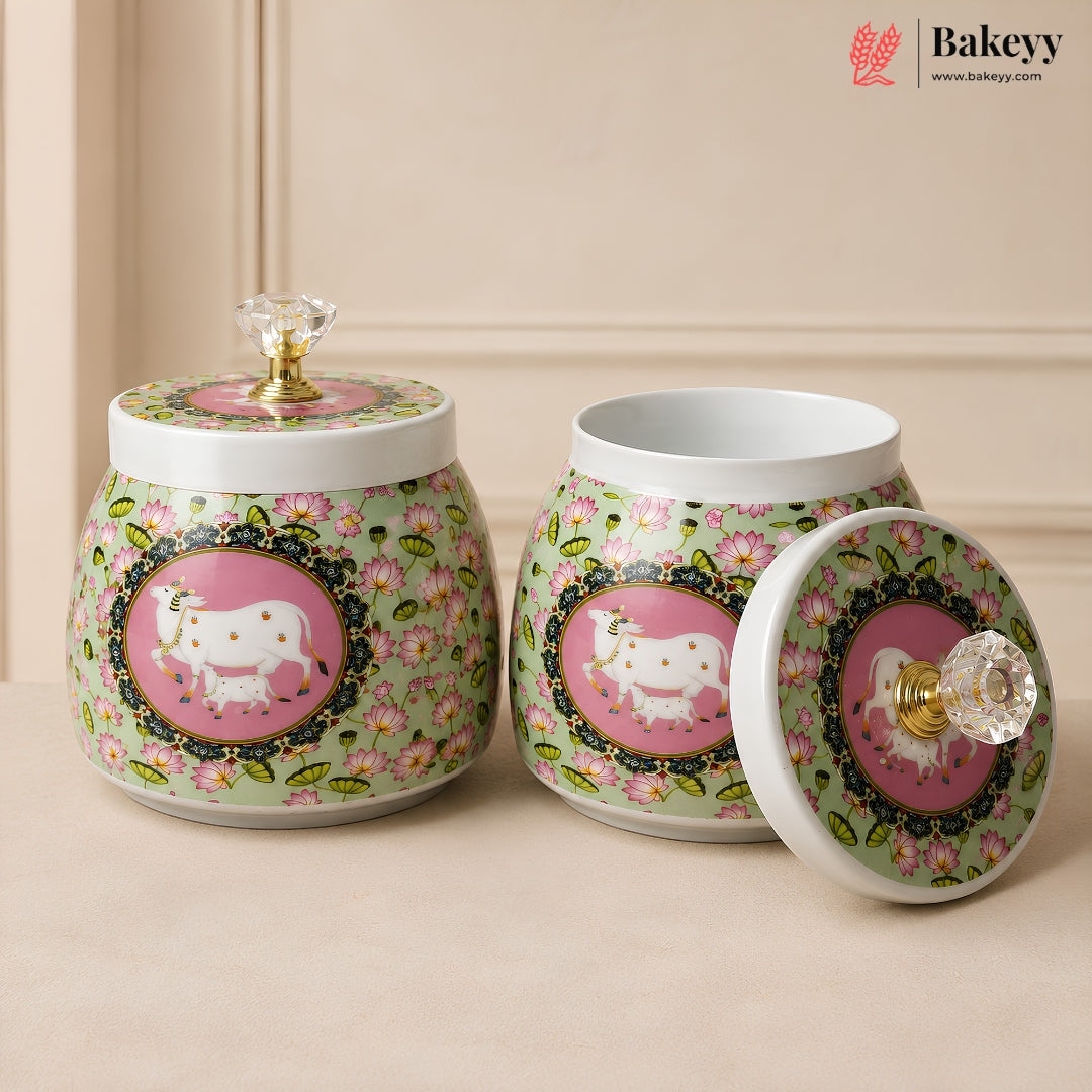 Two decorative ceramic canisters with matching lids, featuring a light green floral background, pink oval panels with white goat motifs, and clear crystal knob handles. One canister is closed and the other is open, set against a warm beige background.