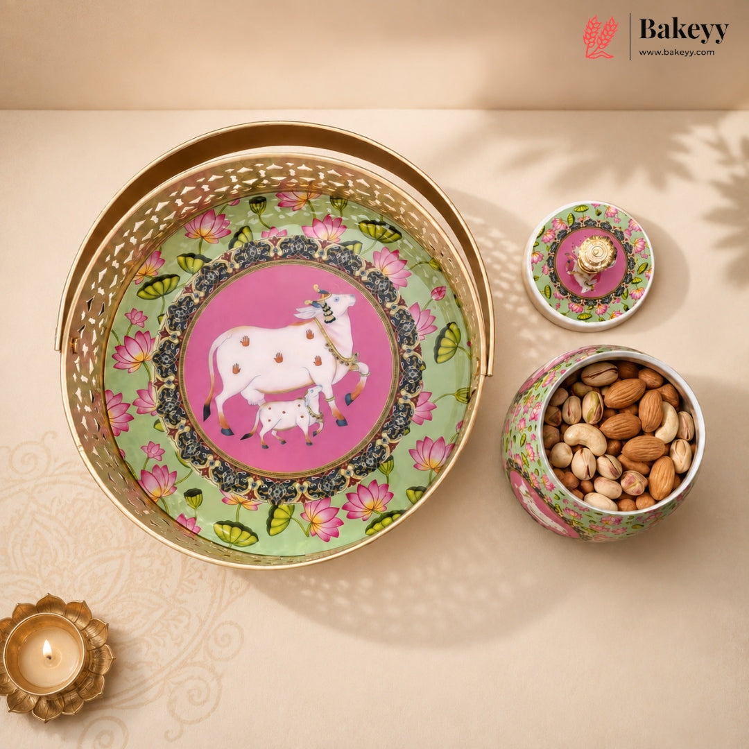 A decorative metal gift set from Bakeyy featuring a large round tray with a pink and green floral design and a white cow illustration, alongside a small lidded tin and an open tin filled with almonds, styled on a beige surface with a lit diya.