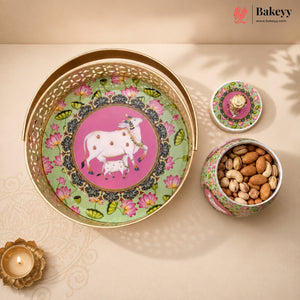 A decorative metal gift set from Bakeyy featuring a large round tray with a pink and green floral design and a white cow illustration, alongside a small lidded tin and an open tin filled with almonds, styled on a beige surface with a lit diya.
