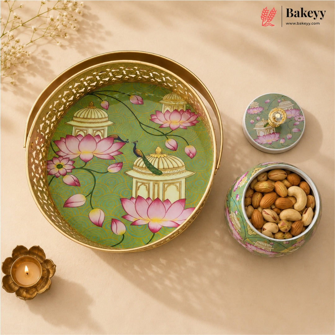 A decorative round metal tray with a gold-tone rim and handle, shown alongside its matching lid and a small open container filled with almonds and cashews. The tray features an illustrated design of pink lotus flowers, domes, and a peacock on a green background.