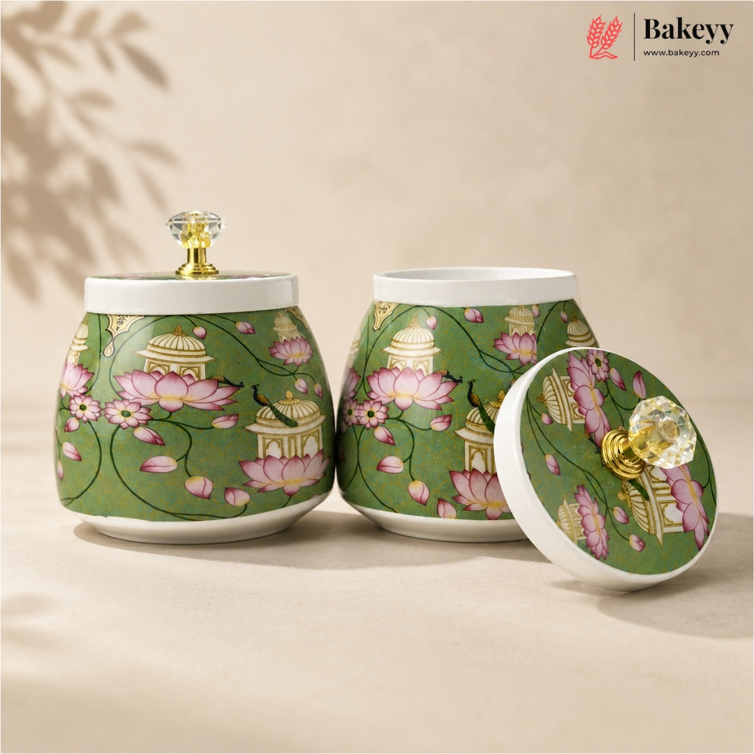 Two green ceramic tea or candy jars with pink lotus and gold architectural floral patterns, shown on a beige background with one jar open and its lid leaning beside it. A Bakeyy logo appears in the top right corner.