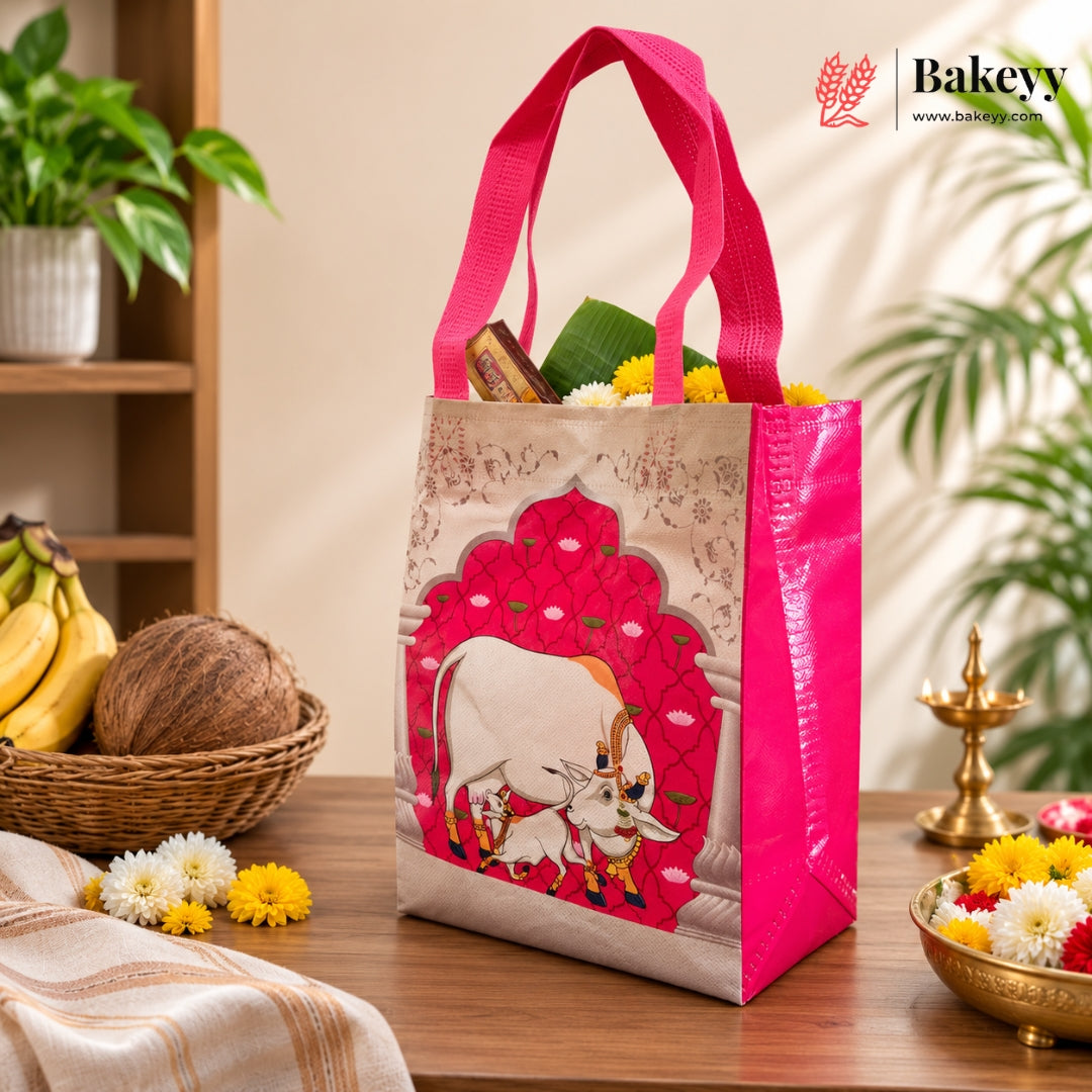 A reusable tote bag with bright pink handles and a pink-and-beige decorative front panel featuring a cow illustration, shown standing on a wooden table in a styled indoor setting with flowers, fruit, and plants.