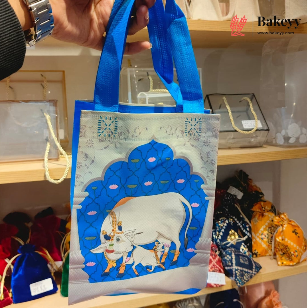 A hand holds a blue reusable tote bag with a printed illustration of a white goat and calf under a blue arch. The bag has blue handles and is shown in a retail setting on wooden shelves with other packaged items in the background.