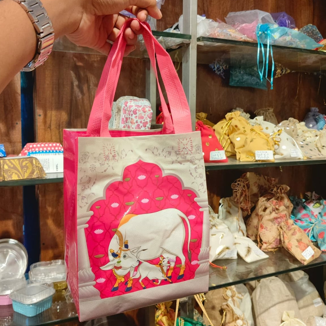 A hand holds a reusable shopping bag with pink handles and a cream-colored body featuring a large pink and white cow illustration. The background shows a retail store shelf filled with various colorful items.