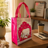 A reusable tote bag with bright pink handles and sides, featuring a printed illustration of a white cow under a red arch on the front, set on a wooden table in a warm indoor environment with a coffee cup, notebook, and plants nearby.