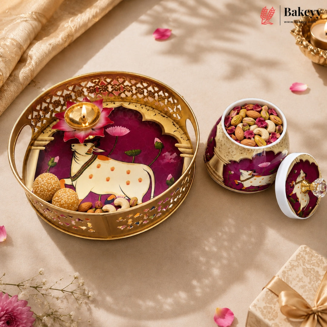A decorative gold-toned tray and matching jar with lid, filled with nuts and dried flowers, styled on a beige surface with scattered petals and a small diya. The Bakey brand logo appears in the top right corner.