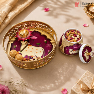 A decorative gold-toned tray and matching jar with lid, filled with nuts and dried flowers, styled on a beige surface with scattered petals and a small diya. The Bakey brand logo appears in the top right corner.