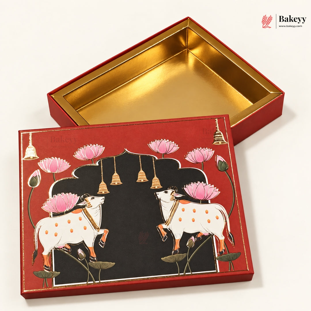A decorative red gift box with a gold interior, shown open with the lid placed beside it. The lid features an illustrated design of two white cows with orange spots under a black arch, surrounded by pink lotus flowers and small gold bells, on a clean white background.