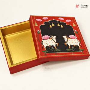 A red and gold square gift box with an illustrated lid featuring two white cows with pink lotus flowers on a black background, shown open on a light beige surface. The interior of the box is gold foil.