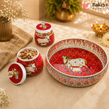 A set of red and white decorative ceramic containers and a matching serving tray with silver-toned embellishments, styled on a beige fabric surface with dried flowers, a brass pot, and a small brass lamp in the background.
