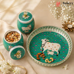 A set of decorative ceramic baking jars and a serving platter in teal green with white cow illustrations, shown on a beige fabric background with flowers and nuts as props. The Bakeyy brand logo is visible in the top right corner.