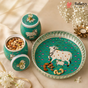 A set of decorative ceramic baking jars and a serving platter in teal green with white cow illustrations, shown on a beige fabric background with flowers and nuts as props. The Bakeyy brand logo is visible in the top right corner.
