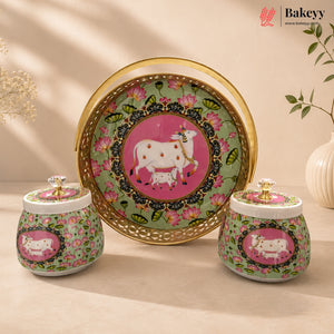 A decorative ceramic set on a beige surface, featuring a large round gold-rimmed tray with a pink cow illustration and two matching lidded jars with floral green patterns and pink cow medallions. The Bakeyy brand logo appears in the top right corner.