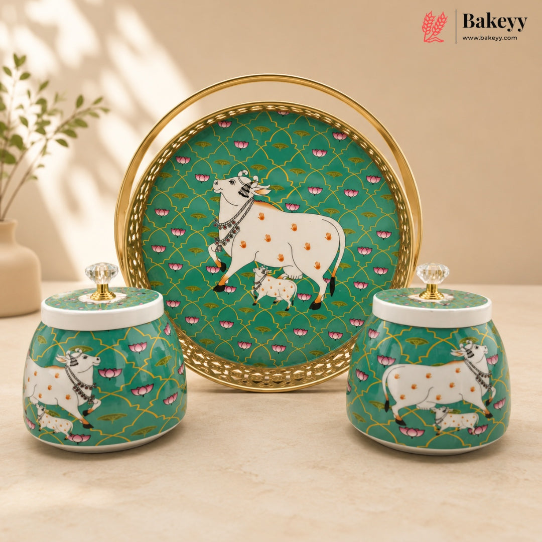 A set of three decorative ceramic items featuring a green background with a gold trellis pattern, white cows and calves, and pink floral accents. The set includes a round serving tray with a gold rim and two matching lidded jars with crystal knobs, displayed on a beige surface with a warm neutral background.