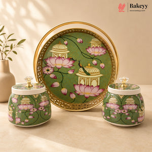 A decorative ceramic baking set by Bakeyy featuring a large round serving tray and two matching lidded jars. The set has a green background decorated with pink lotus flowers, white domed pavilion motifs, and a peacock, framed by a gold-tone border and handle.