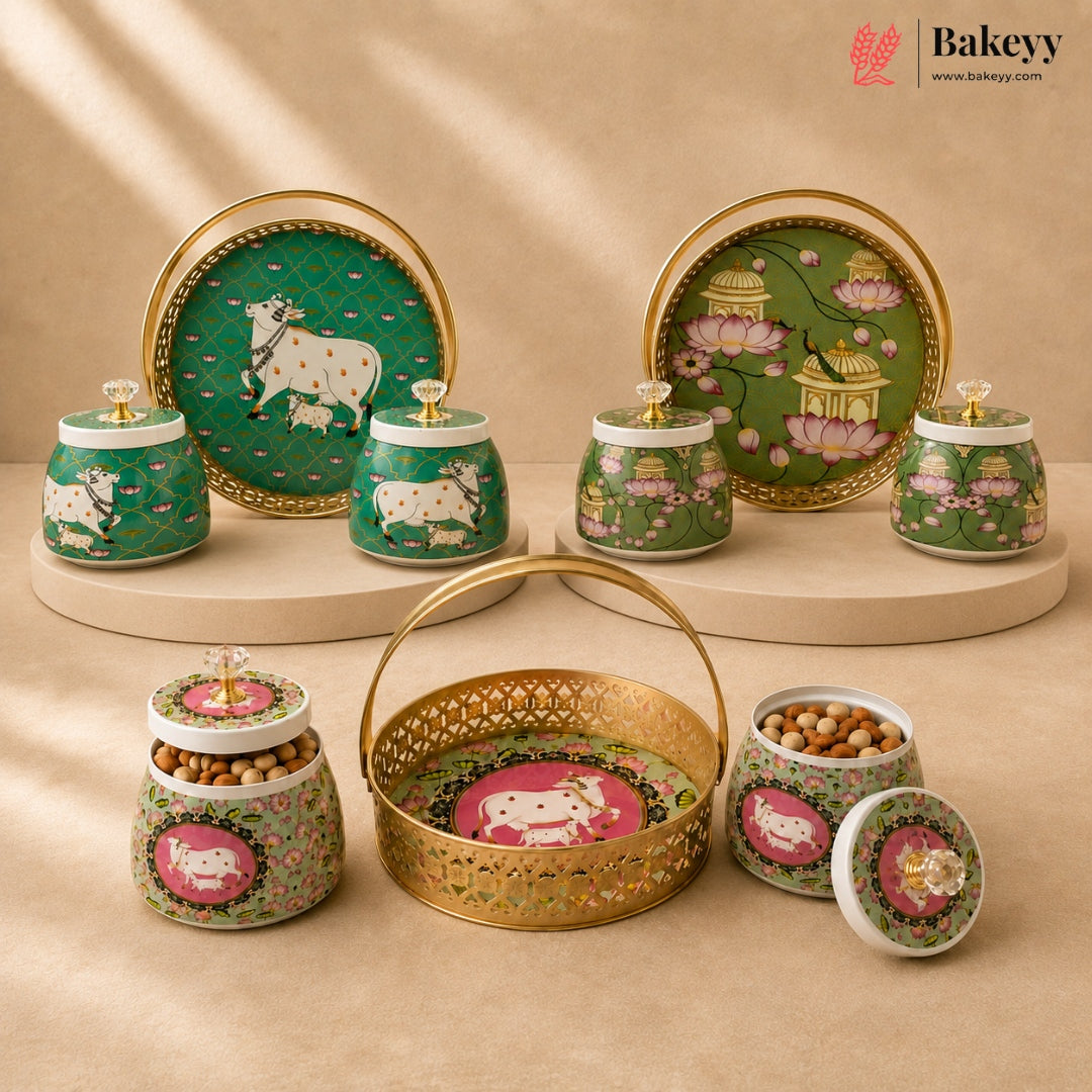 A set of decorative ceramic and metal snack jars, plates, and a basket with ornate green and gold designs, including cow motifs and floral patterns, arranged on beige pedestals against a warm beige background. The Bakeyy brand logo is visible in the top right corner.