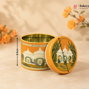 A round metal tin container for baked goods, shown open with its lid leaning against it. The tin has an orange-yellow exterior with a printed illustration of domed architecture and peacocks, and a matching lid featuring a garden scene.