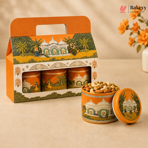 An orange and green illustrated gift box containing three cylindrical tin jars, with one open tin and its lid placed in front, filled with nuts. The packaging features a warm architectural scene with domes and palm trees.