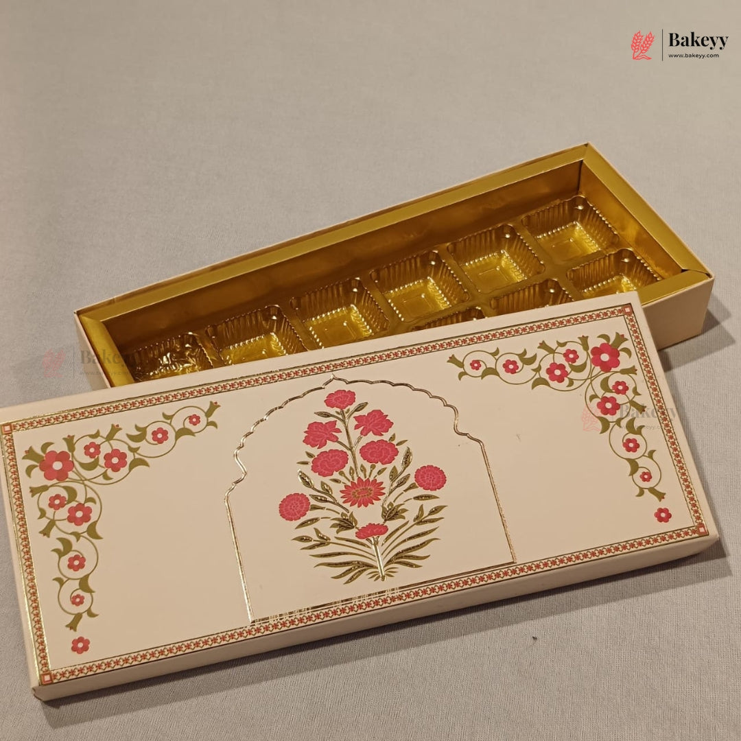 An ornate rectangular gift box for chocolates, shown open with a gold-foil-lined compartmentalized interior. The closed lid in front features cream-colored artwork with pink floral motifs and green vine details.